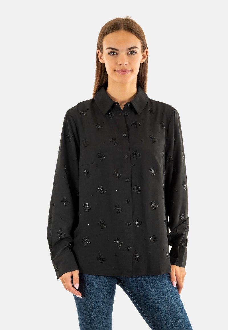 Camicia nera con bottoni e maniche lunghe, caratterizzata da un sottile ricamo floreale nero e una texture liscia. Indossata con jeans in denim blu.