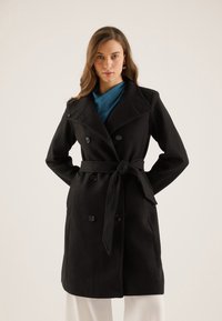 Manteau noir croisé à double boutonnage avec une ceinture à nouer, fabriqué en tissu texturé. Présente un large col à revers et des accents de boutons.