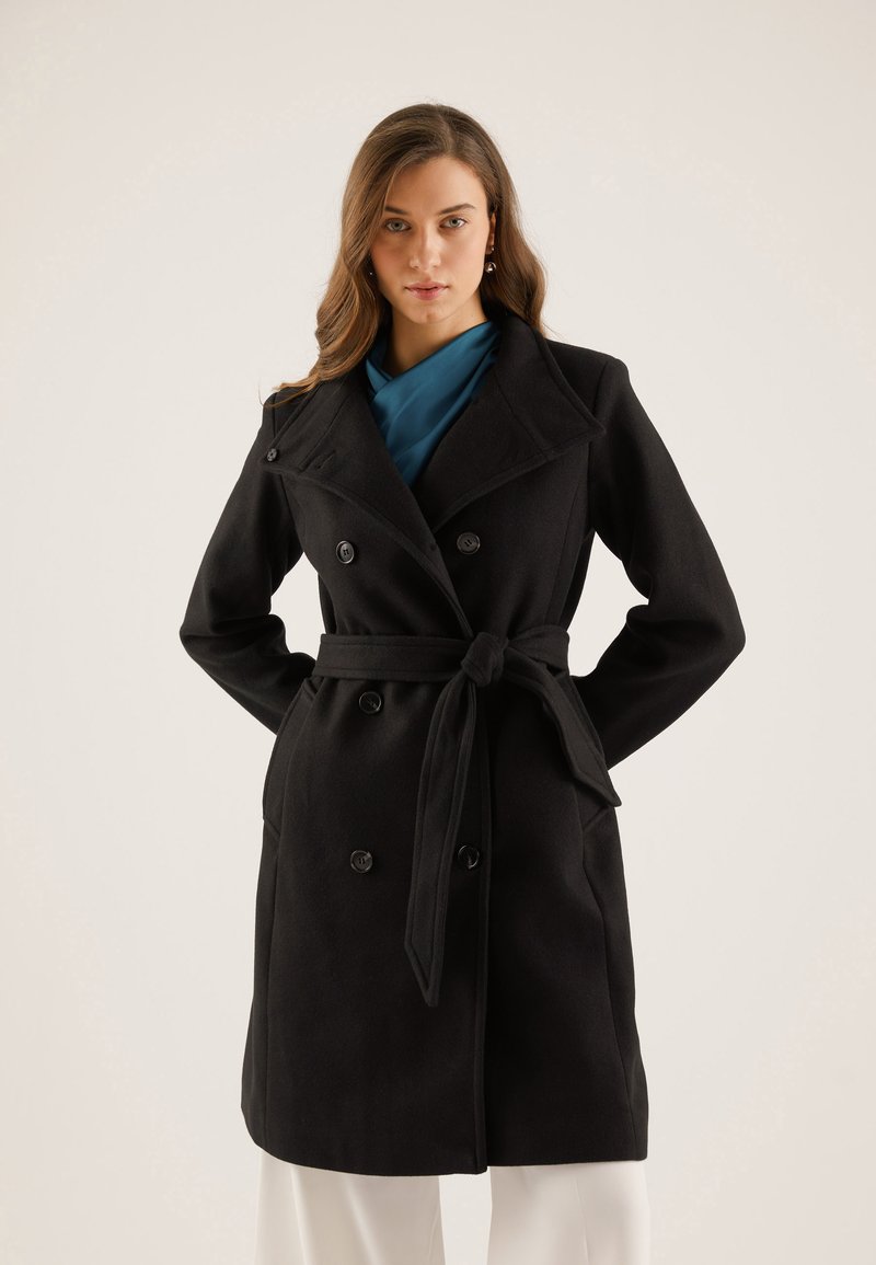 Manteau noir croisé à double boutonnage avec une ceinture à nouer, fabriqué en tissu texturé. Présente un large col à revers et des accents de boutons.