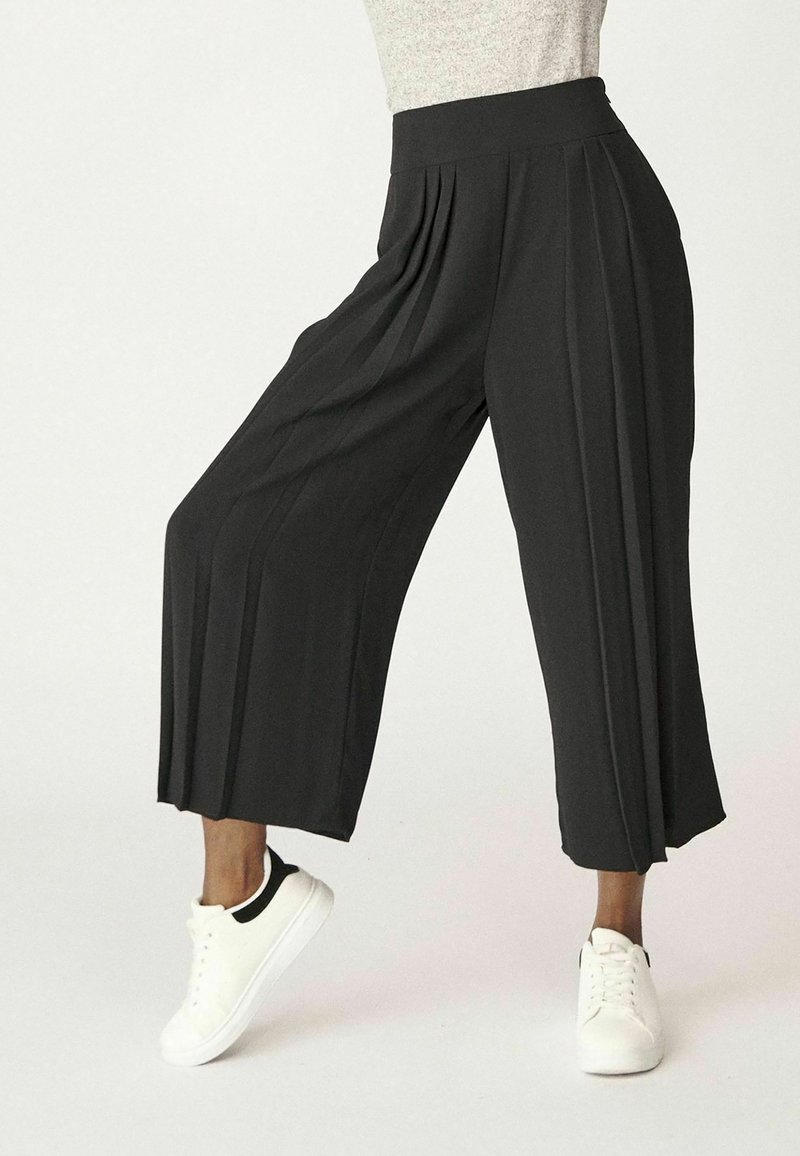 Černé plisované kalhoty z lehkého materiálu s elastickým pasem, spárované s bílými teniskami.