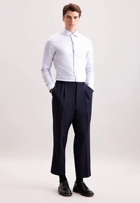 Jasnoniebieska koszula w paski i ciemnogranatowe spodnie plisowane. Koszula ma klasyczny kołnierzyk, a model nosi czarne buty.