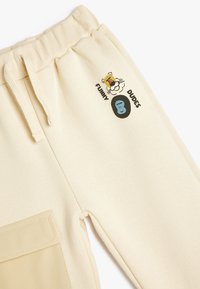 Lys beige sweatpants med snoreliv. Har grafiske prints af tegneserieansigtter og tekst. Inkluderer en sidelomme i en lidt mørkere nuance.