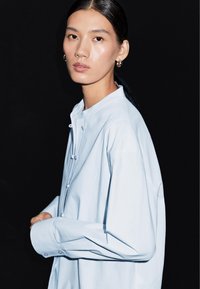 Lichtblauw overhemd met lange mouwen, een mandarijnkraag en knoopdetails. Soepele textuur en licht oversized pasvorm. Model poses tegen een zwarte achtergrond.