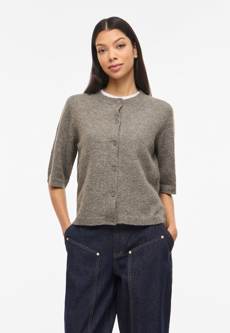 Γυναίκα με μακριά σκούρα μαλλιά, φοράει γκρι κοντομάνικη ζακέτα με κουμπιά και σκούρο μπλε τζιν παντελόνι με φαρδιά μπατζάκια, με τα χέρια στις τσέπες.