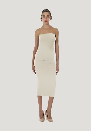 Vrouw met krullend haar en rode lippenstift, die een strapless, getailleerde crèmekleurige midi-jurk en naakte hoge hakken draagt, staand tegen een effen achtergrond.
