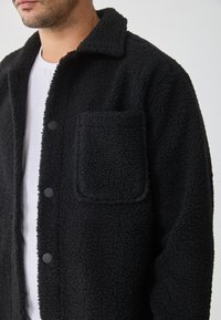 Juodas flisinis švarkas su tekstūruota paviršiumi, turintis apykaklę, mygtukų užsegimus ir krūtinės kišenę.