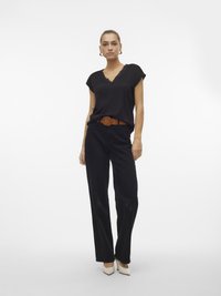 Frau steht vor weißem Hintergrund, trägt ein schwarzes Spitzen-Oberteil mit V-Ausschnitt, schwarze Hose mit braunem Gürtel und weiße spitze Schuhe.