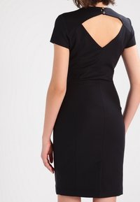 Robe noire ajustée avec des manches courtes, un détail découpé dans le dos et une petite fermeture à bouton au niveau du col. Tissu lisse et forme cintrée.