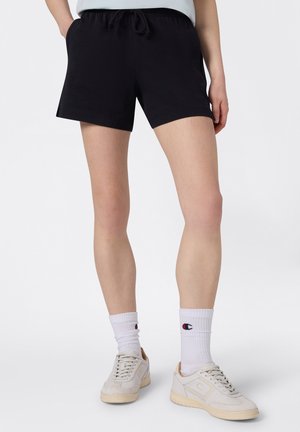 Persona con pantalones cortos negros con cordón, calcetines blancos con el logo de Champion y zapatillas beige, de pie frente a un fondo claro y liso.