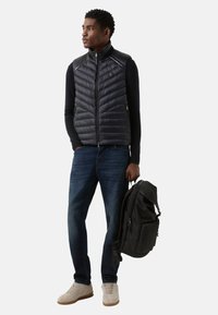 Czarna pikowana kamizelka z zamkiem błyskawicznym z przodu, w połączeniu z ciemnym swetrem i niebieskimi dżinsami. Trzyma czarny plecak, ma na sobie jasne trampki.