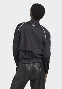 Veste de sport noire avec un design court, accents blancs et taille côtelée. Présente un logo au dos et une texture douce.