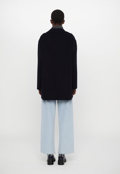 Manteau en laine bleu marine à coupe décontractée, avec des manches larges et un col à revers. Associé à un jean large bleu clair et des chaussures noires brillantes.