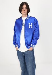 Jachetă bomber din satin albastru cu litera albă 'H', manșete striped și un tricou alb dedesubt, purtată cu blugi din denim albastru deschis.