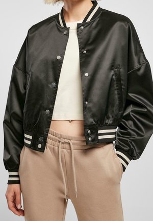 Woman wearing a black satin bomber jacket with white striped cuffs, beige drawstring pants, and a white cropped top, showing a navel piercing.