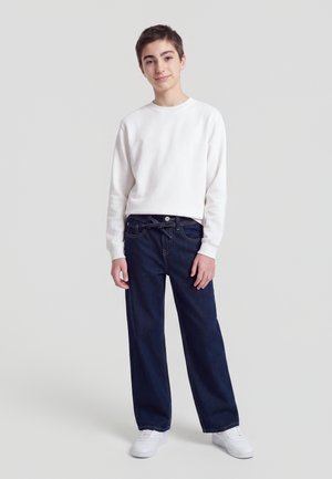 Adolescent debout portant une chemise blanche à manches longues, un jean bleu foncé avec une ceinture nouée, et des baskets blanches sur un fond uni.