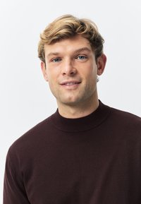 Mørkebrun strikket sweater med høj ribbet hals. Glat tekstur og tætsiddende pasform fremhæver tøjets minimalistiske stil.