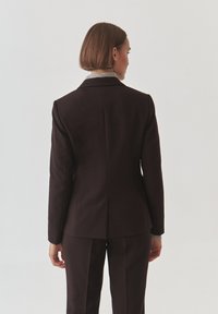 Dark brown tailored suit jacket with sharp lapels, structured shoulders, and a single back vent, paired with matching trousers.