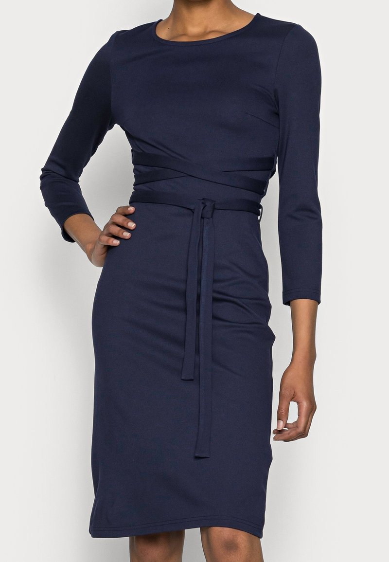 Femme portant une robe bleu marine mi-longue à manches trois-quarts avec une ceinture nouée à la taille, main posée sur la hanche.