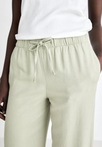 Personne portant un pantalon vert clair à cordon et un haut sans manches en côte blanche, main relâchée sur le côté, sur un fond uni.