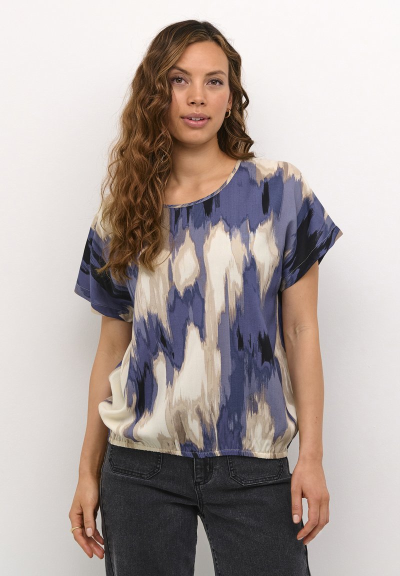 Loose-fitting blouse in blue, cream, and black with a wave pattern. Short sleeves, round neckline, and soft fabric. Paired with dark jeans.