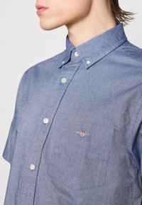 Zbliżenie osoby noszącej niebieską koszulę na guziki z zapiętym kołnierzykiem i kieszenią na piersi z małym wyszywanym logo.