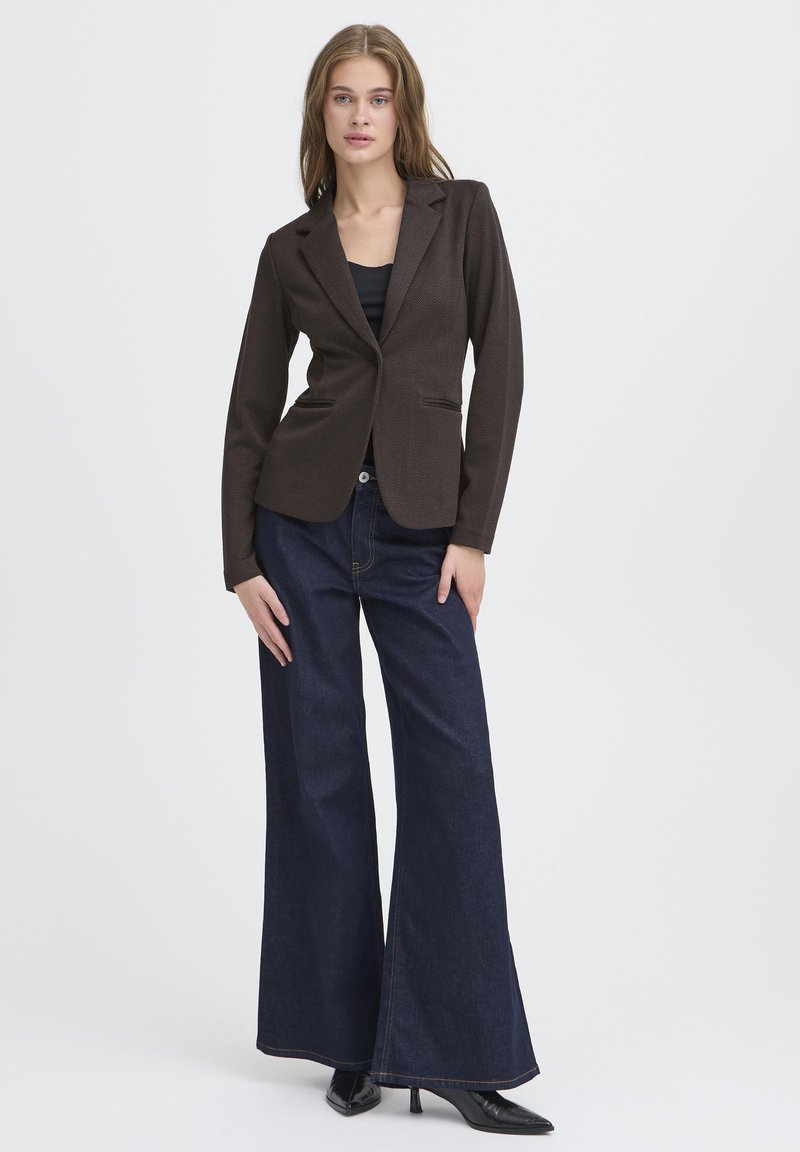 Rudas švarkas su pritaikytu siluetu, dviem priekinėmis kišenėmis ir tekstūruotu paviršiumi, derinamas su aukštu liemeniu tamsiai mėlynomis plačiomis kelnėmis.