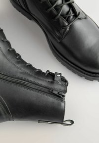 Gli stivali neri in pelle fino alla caviglia presentano design con lacci, cerniere laterali, superficie texturizzata e punta tonda con suola robusta per una buona aderenza.