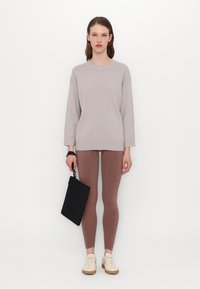Lys grå strikket genser med et teksturert mønster, kombinert med tettsittende brune leggings. Modellen holder en svart clutch og har på seg kremfargede joggesko.