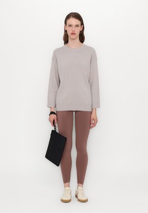 Lys grå strikket genser med et teksturert mønster, kombinert med tettsittende brune leggings. Modellen holder en svart clutch og har på seg kremfargede joggesko.