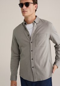 Grijs overhemd met knoopsluiting en een borstzak, gemaakt van een gestructureerde stof, met zwarte knopen en opgerolde mouwen, gecombineerd met een wit T-shirt.