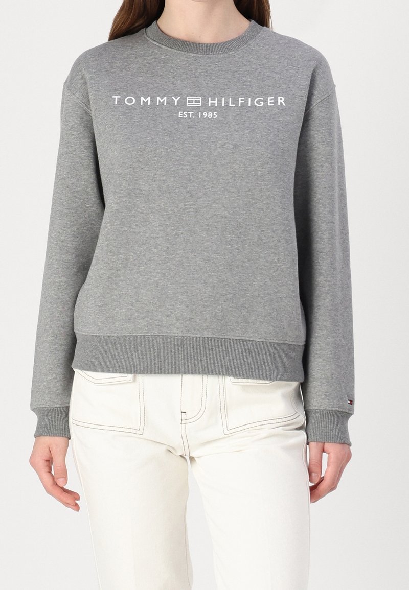 Femme portant un sweat-shirt gris Tommy Hilfiger avec un logo blanc et un pantalon clair sur un fond uni.