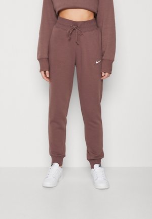 Osoba ubrana w brązowe spodnie dresowe Nike z sznurkiem w pasie i białe sneakersy Nike, stojąca na jednolitym jasnym tle.