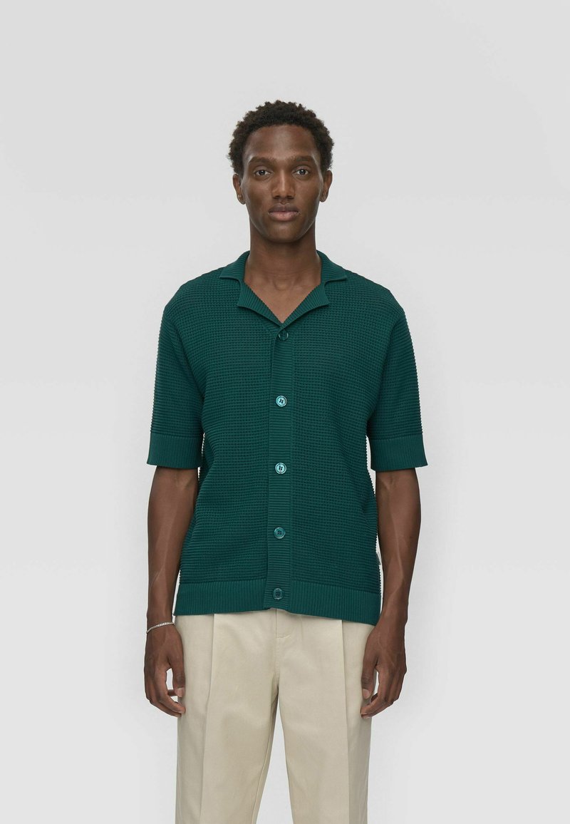 Chemise à manches courtes et à boutons de couleur vert foncé avec un motif texturé et un col, dotée de boutons bleus et de poignets et d'ourlet côtelés.