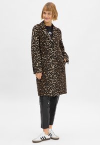Luipaardprint lange jas in warme, geweven stof, met twee zwarte knopen en zijzakken, gecombineerd met zwarte leren broek en witte sneakers.
