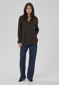 Braun gerippte Hemdbluse mit Kragen und langen Ärmeln, kombiniert mit dunkelblauen weit geschnittenen Jeans und schwarzen spitzen Absätzen.