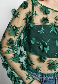 Grünes transparentes Top mit floralen Stickereien, das mit strukturierten grünen Applikationen an Ärmeln und Oberteil versehen ist, kombiniert mit Jeans.