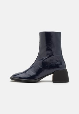Lesklé námořnicky modré kotníkové boty s špičatou špičkou, středně vysokým blokovým podpatkem a elegantním designem. Vyrobeno z lakovaného materiálu s hladkým povrchem.