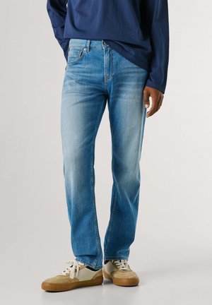 Uomo che indossa jeans azzurri slavati, sneakers beige e bianche e una maglia blu navy a maniche lunghe, in piedi davanti a uno sfondo semplice.