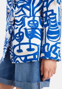 Weiße Jacke mit blauen abstrakten Mustern, ausgestattet mit einer einzelnen Tasche. Kombiniert mit Jeans-Shorts mit fransigem Saum.