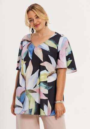 Vrouw met blond haar, gekleed in een zwarte blouse met bloemenprint, V-hals en wijde korte mouwen, en lichtgekleurde broek, staand tegen een effen achtergrond.