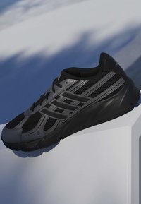 Sort og grå atletisk sneaker med mesh og syntetiske overlays, med en tekstureret sål, hvilende på en hvid overflade med blå baggrundsskygger.