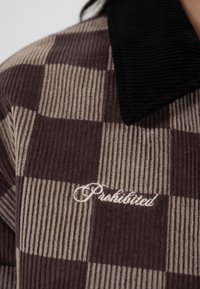 Gros plan sur une veste en velours côtelé à carreaux marron et beige avec un col noir et le mot brodé "Interdit" en blanc sur la poitrine.