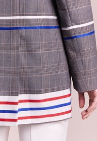 Blazer grigio a quadri con strisce blu, bianche e rosse lungo l'orlo inferiore, caratterizzato da una forma su misura e un tessuto testurizzato.