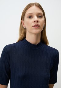 Navyblauwe, textuurstof top met hoge hals en korte mouwen. Kenmerkt zich door een geometrisch patroon en is gemaakt van een lichtgewicht stof. Zilveren oorbellen inbegrepen.