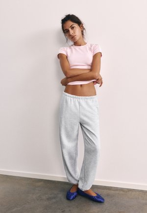 Junge Frau trägt ein hellrosa Crop-Top, graue Jogginghose und blaue Ballettschuhe, steht mit verschränkten Armen vor einer schlichten Wand.