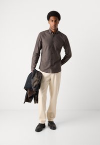 Cămașă gri cu nasturi, pantaloni de culoare crem, mocasini negri; ținând o jachetă din denim închis; material neted, design simplu și croială ajustată.