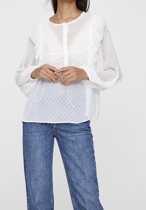 Femme portant un chemisier blanc transparent à pois avec des volants et des manches longues, associé à un jean en denim bleu, mains croisées à la taille.