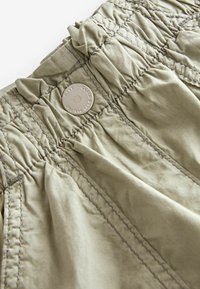 Khaki tygmidja med samlad textur, med en silvermetallic knapp och kontrasterande sömnad. Närbild av detalj.