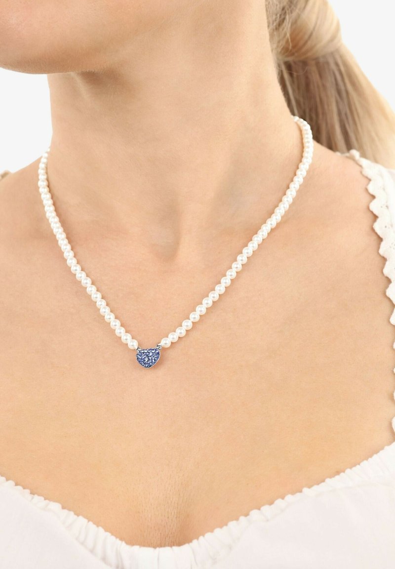 Donna che indossa una collana di perle con un piccolo ciondolo a forma di cuore in pietra blu, vestita con un top bianco a balze.