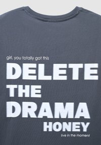 Maglietta grigia con una scritta bianca in grassetto che dice "ELIMINA IL DRAMA TESORO" e piccole scritte "ragazza, ce la fai alla grande" e "vivi nel presente."